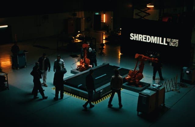 The image depicts a warehouse setting with a group of people gathered around a large, dark gray or black robotic arm. The robotic arm is positioned on a raised platform with a yellow and black striped safety line. The warehouse has a dark, industrial aesthetic with a few pieces of equipment visible in the background. A