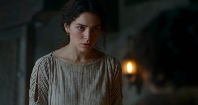 The image shows a woman with dark, curly hair wearing a beige, textured top. She is standing in a dimly lit room, with a blurred background that suggests a stone or brick wall. The woman's gaze is directed off to the side, and her expression appears serious or concerned. The lighting in the room is dim, with a warm, ye