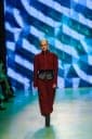 The image captures a fashion show scene. A model is the central focus, striding confidently down a runway. She is wearing a vibrant red jumpsuit with a contrasting black belt and black boots. The runway is a striking blue, with a geometric pattern of white and light blue triangles. The background features a large scree