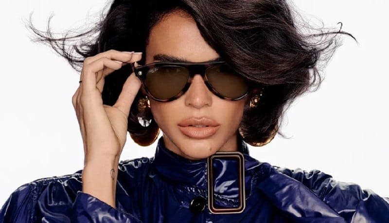 The image shows a person with dark brown or black hair, wearing a navy blue jacket with a shiny, possibly patent leather, collar. The jacket has a distinctive gold-colored rectangular detail on the front. The person is also wearing large, dark brown or black sunglasses with a tortoiseshell or brown and black pattern. T
