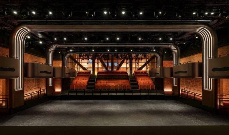 The image depicts a grand theater with a high ceiling and a black stage. The stage is flanked by rows of orange seats, and the walls are a combination of brick and a dark material, possibly metal or dark wood. The theater features a curved, metallic-looking structure above the stage, and the stage is lit by spotlights.