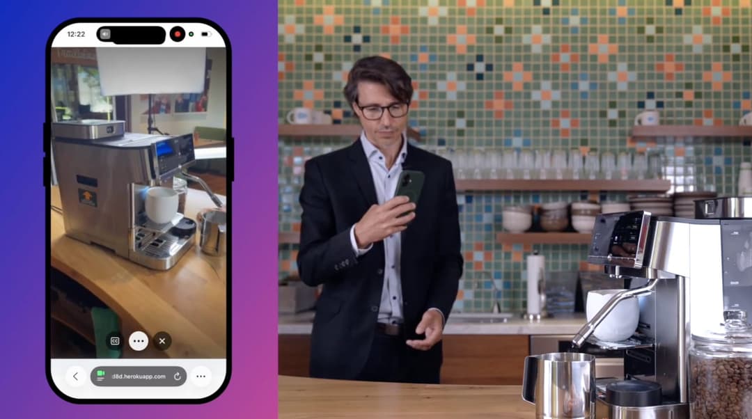 The image shows a man in a black suit and glasses standing in front of a coffee machine in a kitchen. He is holding a smartphone in his right hand and appears to be looking at it. The coffee machine is a modern, stainless steel model with a digital display. The kitchen features a green and orange tiled backsplash and a