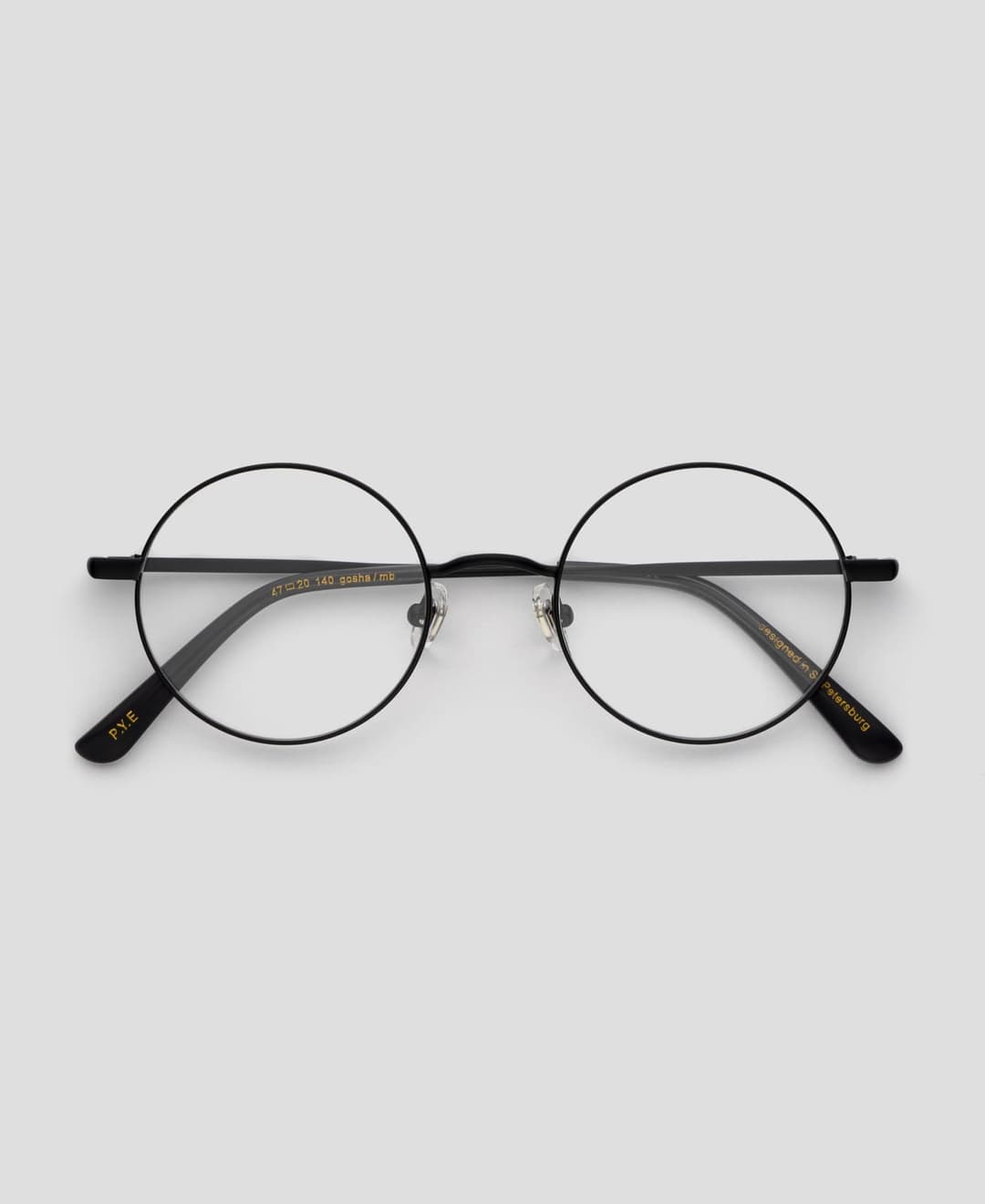 The image shows a pair of black-framed round glasses resting on a light gray background. The glasses have thin black arms and a thin black frame. The lenses are clear, allowing the light to pass through. The glasses are positioned with the temples slightly angled upwards. The brand name "P.Y.E" is visible on the left t