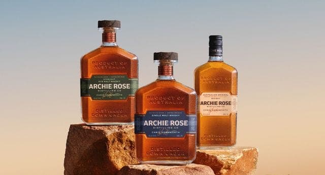 The image shows three bottles of "Archie Rose" whiskey, each with a distinct label design, arranged on a rock formation. The bottles are a golden-brown color, and the labels feature a combination of green and blue colors. The leftmost bottle has a green label with the words "Product of Australia" and "Archie Rose", whi