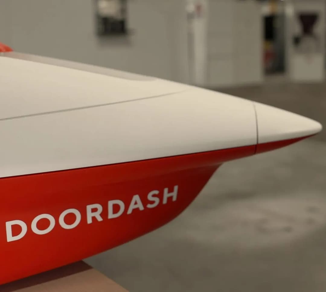 The image shows a close-up view of a model of a DoorDash delivery vehicle. The model is predominantly red and white, with the word "DOORDASH" prominently displayed in white letters on the side. The model is positioned on a surface that appears to be a table or platform. The background is blurred, suggesting an indoor s
