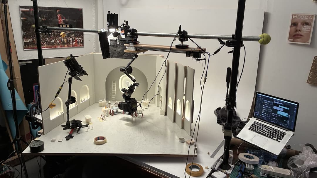 The image captures a scene from a film or television set production, featuring a white wooden set with a black camera mounted on a tripod. The set is illuminated by a combination of natural light and artificial lighting, with a large black light source positioned on the left side. The set is surrounded by a black metal