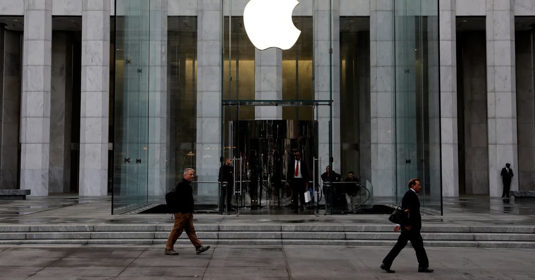The image shows the entrance to the Apple Inc. building, a modern architectural structure with a glass facade. The entrance features a large, white Apple logo prominently displayed above the glass doors. The building is constructed of light gray or beige stone, and the entrance is framed by a series of columns. Several