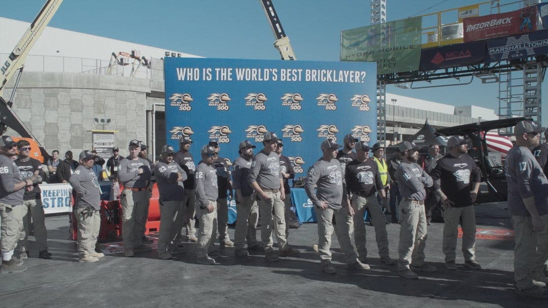 The image shows a group of people standing in front of a large blue banner that reads "Who is the world's best bricklayer?". The banner features a repeating pattern of the word "Bricklayer" and the number "500". The group is dressed in gray t-shirts with a logo and khaki pants, and they appear to be participating in a
