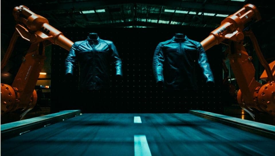 The image shows two robotic arms, one on each side of a black mannequin torso, suspended in a warehouse setting. The mannequin is wearing a black leather jacket. The warehouse has a dark, industrial aesthetic with a black wall featuring a grid pattern. The floor is a dark blue or teal color, and a white line runs down