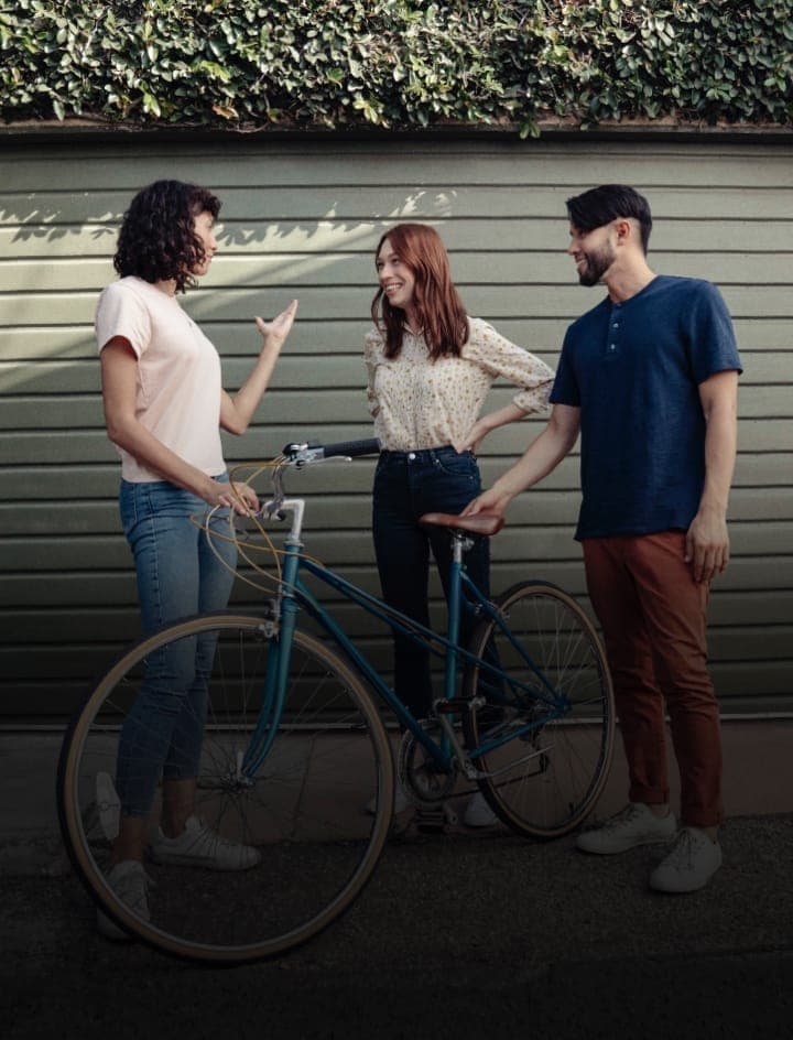 The image shows three individuals standing in front of a green wall with a horizontal siding pattern. The wall is adorned with a row of green bushes or shrubs. On the left, a woman with curly hair is engaged in a conversation with two men. She is wearing a light pink/beige t-shirt and blue jeans. The man on the right i