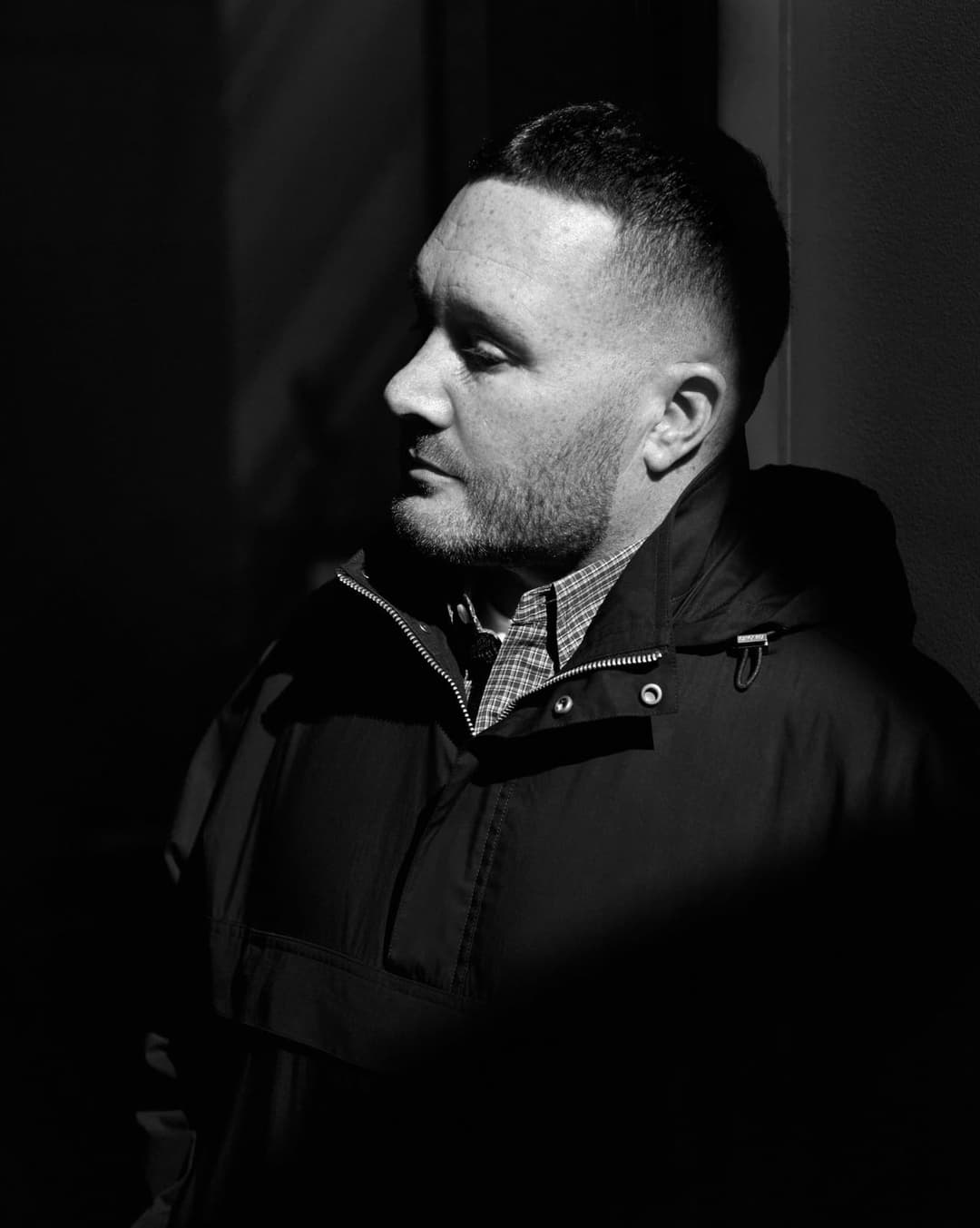 This black and white photograph captures a man in profile, standing against a dark background. He is wearing a dark jacket with a zipper and a collared shirt underneath. The man's gaze is directed to the left, and his facial expression is neutral. The lighting in the photograph is dramatic, with a light source illumina