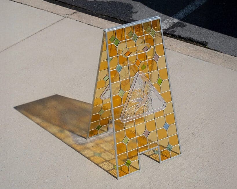 The image shows a vibrant yellow caution sign with a diamond pattern and a white triangle with a black figure, possibly a person, on it, standing upright on a light beige sidewalk. The sign is made of stained glass with a mix of yellow, orange, and green hues. The sign is positioned slightly off-center to the left, cas