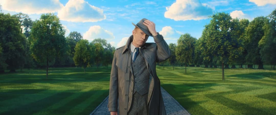 The image shows a man standing on a path in a park. He is dressed in a gray suit and a gray fedora, and is holding his hat in his right hand. The path he stands on is made of cobblestones or pavers. The park around him is lush and green, with trees lining the path. The sky above is a clear blue with fluffy white clouds