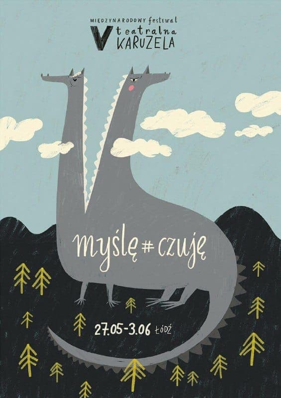 The image is a poster for the Miedzynarodowy festival, featuring a stylized illustration of two gray dragons with white teeth and horns. The dragons are positioned on a dark gray/black hill, with a few stylized trees scattered around them. The background is a light blue sky with white clouds. The text on the poster inc