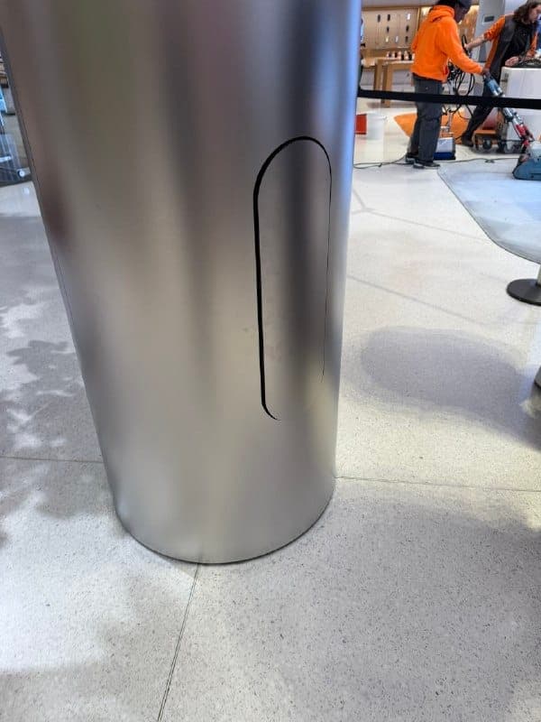 The image shows a large, cylindrical, metallic object, possibly a part of a display or kiosk, with a black, oval-shaped cutout on its side. The object is situated on a light gray floor with a speckled pattern. In the background, a person wearing an orange jacket is visible, and a portion of a second person is also seen