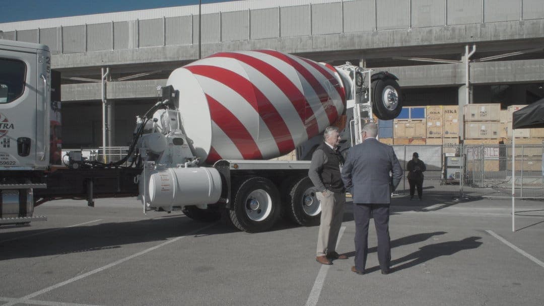 The image shows a large concrete mixer truck with a red and white striped body parked in a parking lot. The truck has a large red and white striped container on its back, and a white tank is attached to the side. Two men are standing near the truck, engaged in conversation. One man is wearing a dark suit and the other
