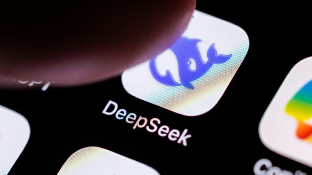 The image shows a close-up view of a smartphone screen displaying the app icon for "DeepSeek". The app icon is a blue dolphin, outlined in white, set against a white background with a subtle rainbow-like gradient. The word "DeepSeek" is written in white text below the icon. The phone's screen is black, and a finger is