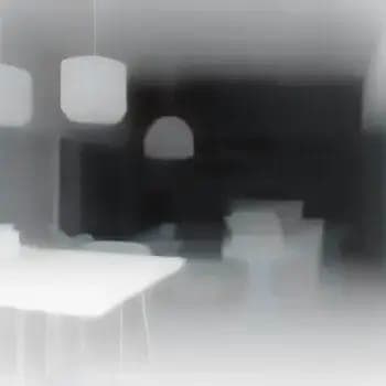 The image presents a modern interior space, captured in a black and white photograph. The room is dominated by a white table, which appears to be the central focus. The table is surrounded by a few chairs, though their details are obscured by the soft blur. The room is illuminated by a hanging lamp, which casts a soft