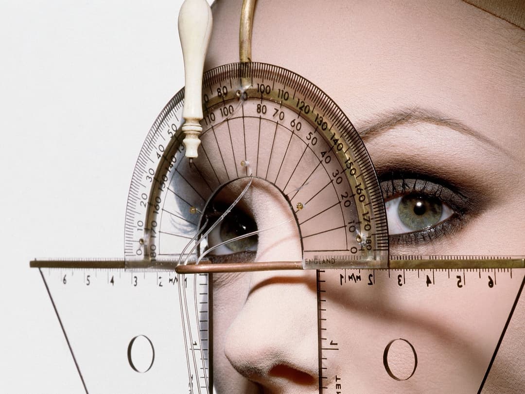 The image shows a close-up view of a person's face, with the nose and eye area prominently featured. The person's eyes are open, and they are wearing makeup, including eyeliner and eyeshadow. A gold-colored metal rod with a white handle is positioned above the person's head, holding a clear plastic or glass measuring t