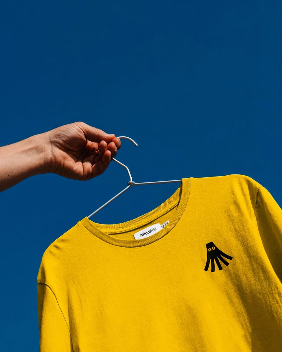 The image shows a hand holding a white clothes hanger against a clear blue sky. The hanger is suspended from the hanger bar, which is attached to a yellow t-shirt. The t-shirt features a black octopus design on the left chest area. The t-shirt has a round neckline and a crew neck. The hand holding the hanger is positio