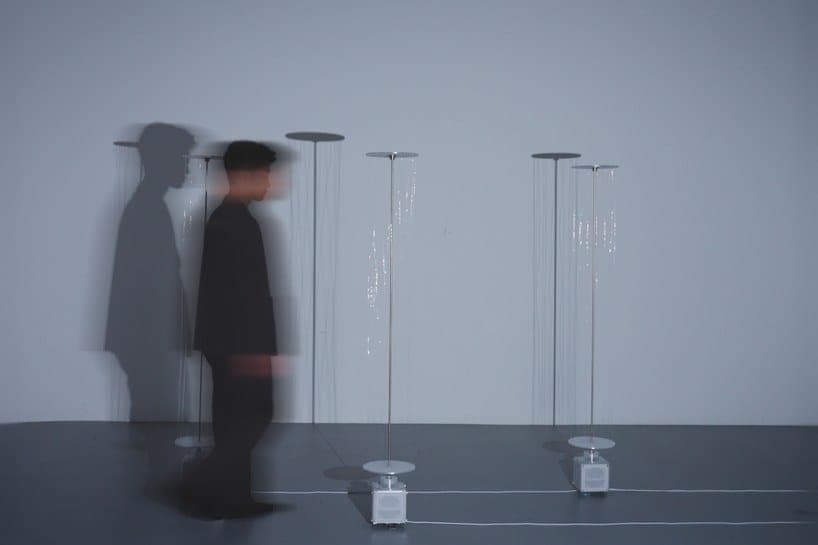 The image shows a minimalist art installation featuring three tall, slender, white lamps with circular bases. The lamps are arranged in a loose triangular formation against a light gray wall. The lamps are illuminated from within, casting a soft glow. The floor is a dark gray or black, and the overall color palette is