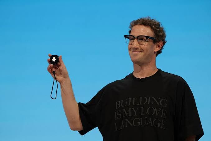 The image shows a man standing against a vibrant blue backdrop. He is wearing a black t-shirt with the phrase "Building is my love language" printed on it in white letters. The man is holding a small, black, round object in his right hand, which appears to be a smartwatch or similar device. He is also wearing glasses a
