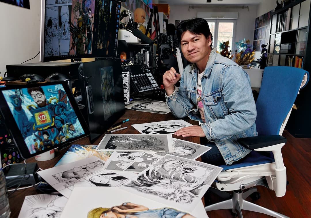 The image shows a man seated at a desk in a home office, engrossed in his work. He is wearing a light blue denim jacket and has dark hair. The desk is cluttered with a variety of papers and drawings, including black and white sketches, and a computer monitor displaying a comic book-style image. The man is holding a pen