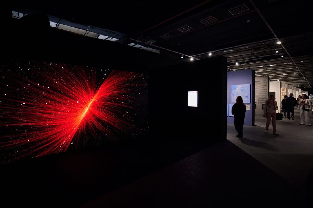 The image depicts a modern art gallery with a large, dark-colored screen displaying a vibrant red and orange abstract design, possibly a representation of a starburst or cosmic event. The room is dimly lit, with spotlights illuminating the screen and the dark floor. Two individuals are standing near the screen, one wea