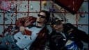 The image shows two men lying on a tiled floor, possibly in a room or hallway. The man on the left is wearing a red jacket and sunglasses, holding a red object, possibly a phone. The man on the right is wearing a black jacket and a baseball cap, and is holding a bag of popcorn. The floor is covered in scattered red and
