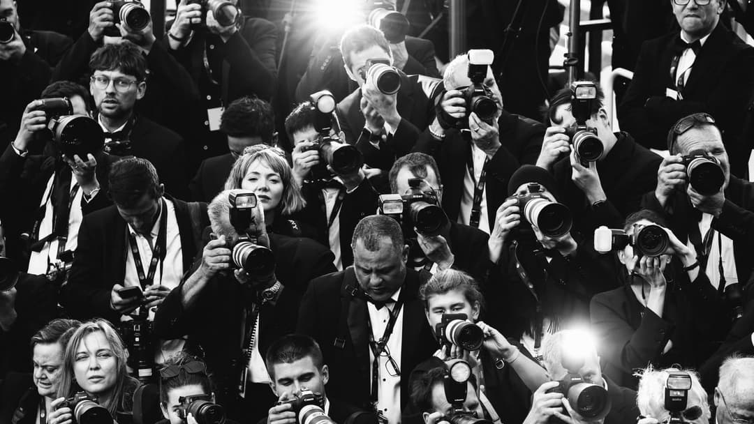 This black and white photograph captures a group of people, primarily men, dressed in formal attire, including suits and ties, standing in a line. Each person is holding a camera, suggesting they are photographers or reporters documenting an event. The people are arranged in a somewhat staggered formation, with some st