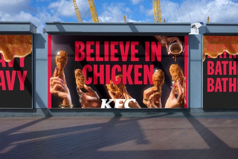 The image shows a large billboard advertisement for KFC, located in London, UK. The billboard features a vibrant red and black color scheme with the text "Believe in chicken" prominently displayed in bold white letters. The billboard also includes a collage of images of hands holding and eating fried chicken pieces, wi