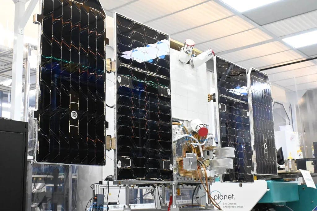 A satellite with dark deployed solar panels and a robotic arm is displayed in a bright cleanroom, with a "planet." logo and the tagline "See Change. Change the World." on its base.