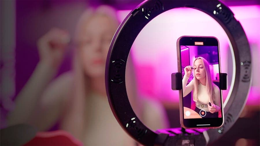 The image shows a person with blonde hair, wearing a white shirt, using a makeup application tool, possibly a lip gloss or lipstick, on their face. The person is positioned in front of a black ring light with a circular design, which is attached to a smartphone. The smartphone screen displays a video call with the pers