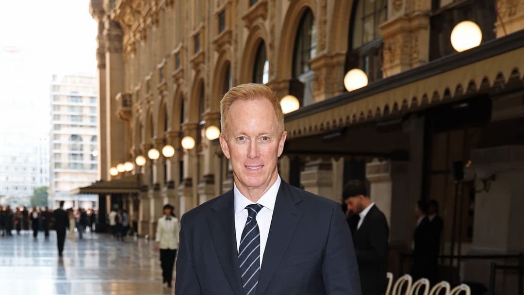 The image shows a man in a dark blue suit and striped tie standing in front of a grand, ornate building with a beige facade and arched windows. The building has a decorative awning or overhang. The man is looking directly at the camera with a slight smile. The building is situated in a bustling city, with other people
