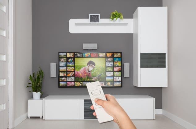 A hand holds a white remote control pointed at a wall-mounted flat-screen TV showing a grid of video thumbnails and a child in a pink top, in a modern living room with a gray wall, white cabinets, a potted plant, and a wall clock. No brand is visible.