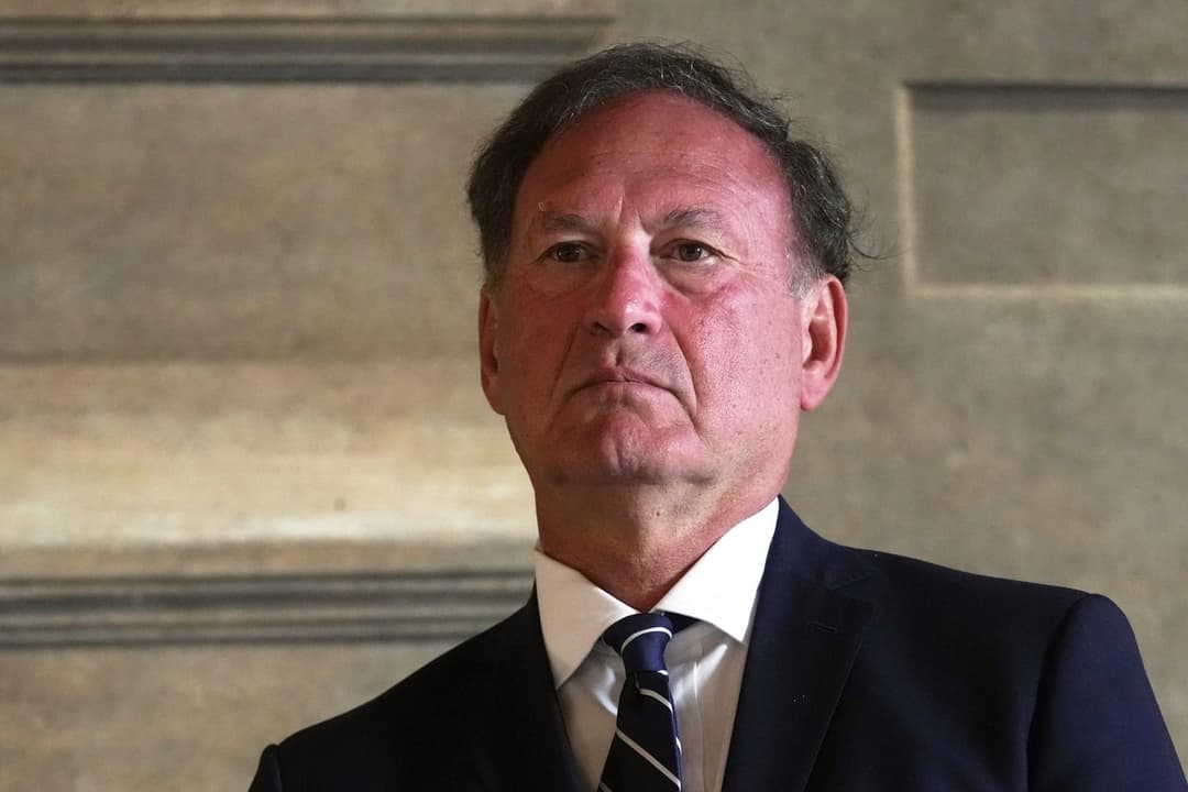 The image shows a man in a suit and tie, standing against a textured, light-colored stone wall. The man is wearing a dark suit and a white shirt, with a dark blue and white striped tie. His hair is dark and appears to be styled in a somewhat tousled manner. His gaze is directed off to the side, and his expression is se