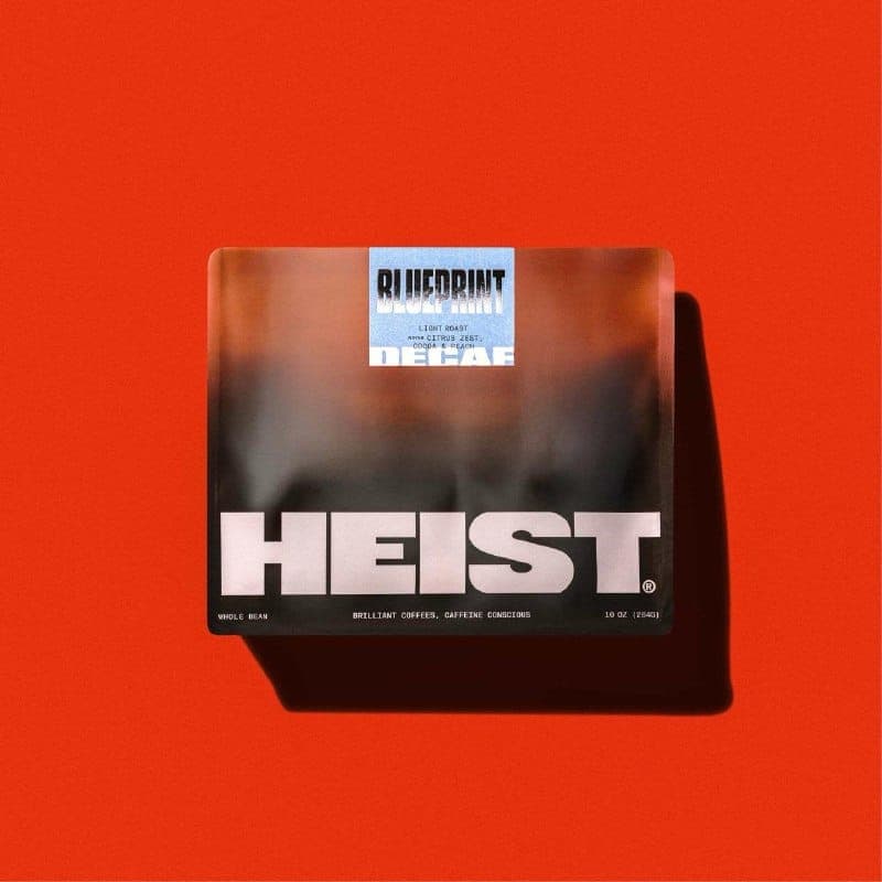 The image shows a rectangular box of "Heist" coffee, set against a vibrant red background. The box is primarily black with a gradient effect transitioning from dark brown to a lighter brown. The brand name "Heist" is prominently displayed in bold white letters, with the tagline "Light Roast" and "Coffee & Peach" writte