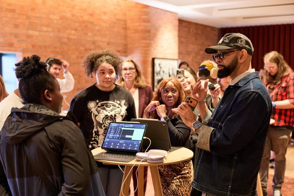 The image shows a group of people gathered around a table in a room with exposed brick walls and a red curtain. The table holds two laptops, one open and displaying a music production software interface, and a small white device, possibly a MIDI controller. A man in a dark blue denim jacket and a baseball cap is holdin
