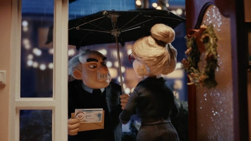 The image shows a man and a woman standing in front of a window, with a wreath hanging on the window frame. The man is holding a black umbrella and a brown envelope, while the woman is holding a black umbrella. The scene is set against a backdrop of a city street at night, with lights visible in the background. The man
