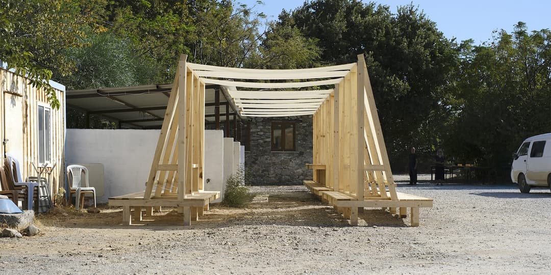 The image shows a wooden structure, possibly a temporary shelter or awning, constructed from light-colored wooden beams and supported by sturdy posts. It is situated in a gravel area, with a small, light-colored building with a corrugated metal roof and a white wall nearby. A white van is parked in the background. The
