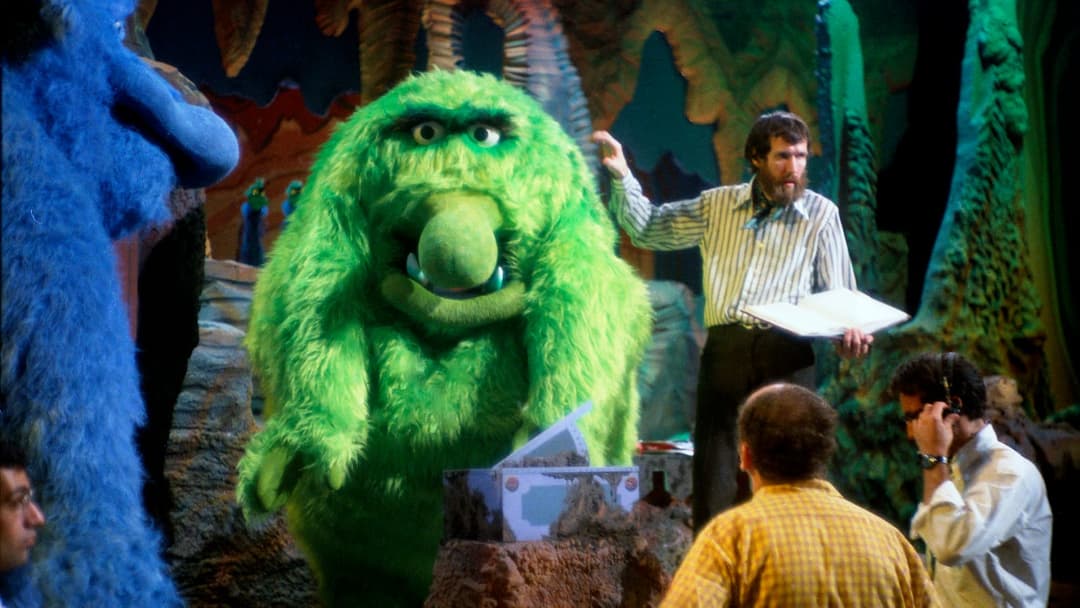 The image captures a scene from a stage performance, featuring a large, green, furry monster as the central focus. The monster has a wide, friendly smile and is standing on a rock formation. To the left of the monster, a blue, furry character is visible. On the right side of the stage, a man in a striped shirt is holdi