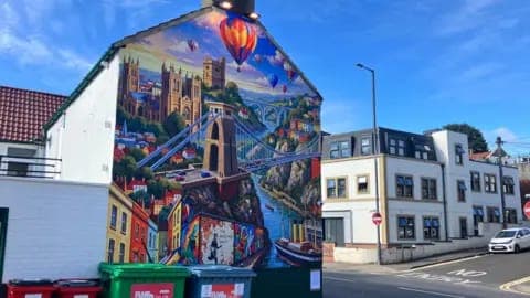 The image depicts a vibrant mural painted on the side of a white building, situated on a street corner. The mural features a colorful depiction of a cityscape with a prominent bridge, a castle, and a hot air balloon, along with a river or canal. The mural is set against a backdrop of a clear blue sky. The street in fro