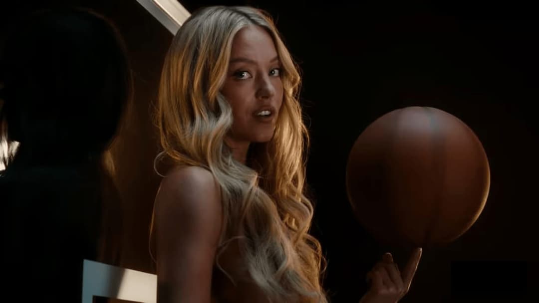 A woman with long, wavy blonde hair looks back over her shoulder in a dark room while holding a large orange-brown spherical object on her fingertip. The image has no visible text or brand marks.