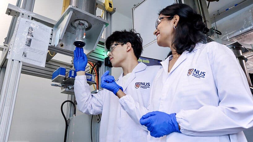 The image shows two individuals in white lab coats and blue gloves standing in front of a large, metallic machine with a complex structure. The machine appears to be a piece of laboratory equipment, possibly a spectrometer or analyzer, with a blue light source at its top. One person is holding a small, dark object, pos