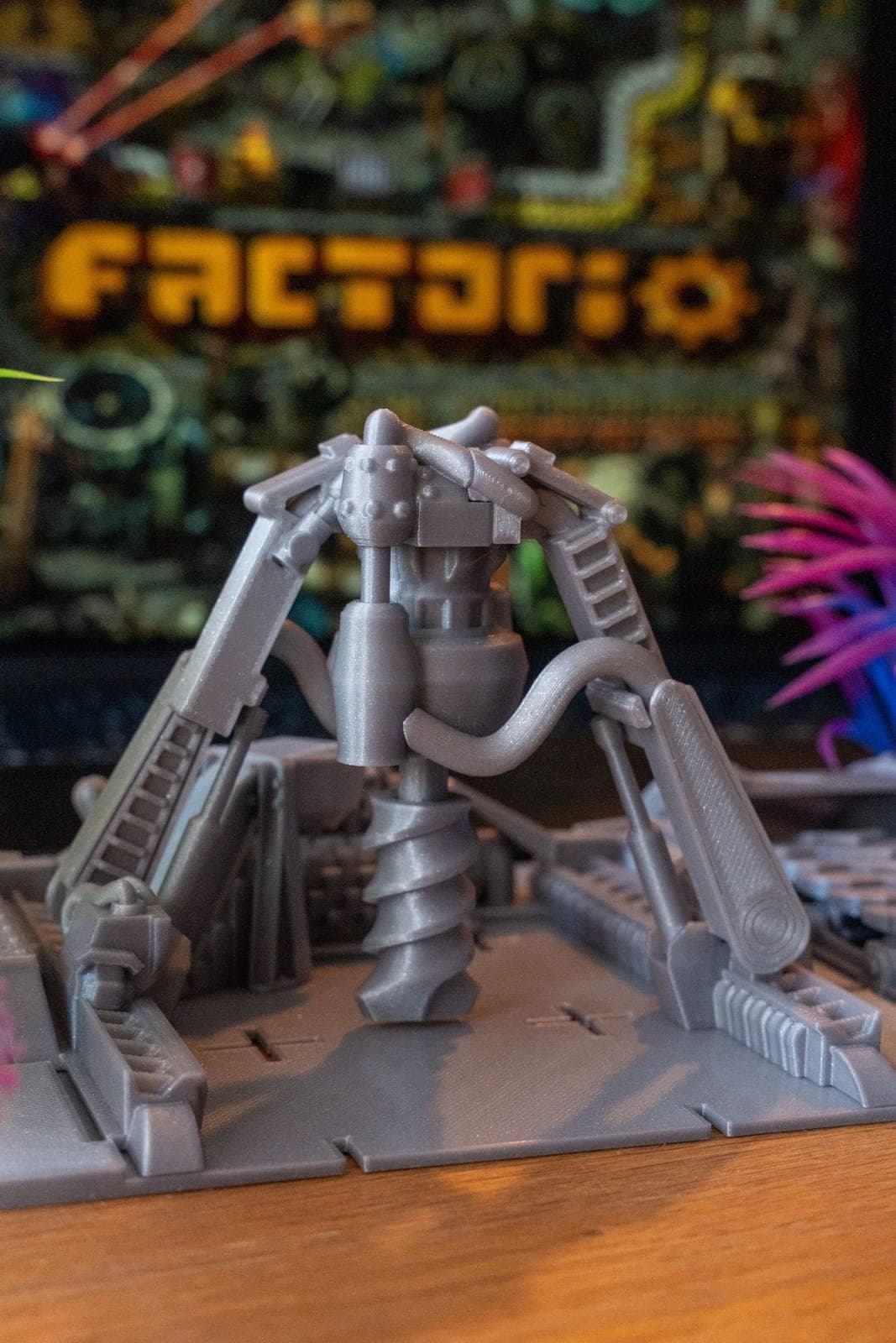 The image shows a gray metal robot figurine standing on a gray base. The robot has a complex design with various mechanical components, including a large head, a body with a cylindrical shape, and a claw-like hand. The base features a spiral-shaped structure, possibly a drill bit, and a set of stairs or steps. The robo