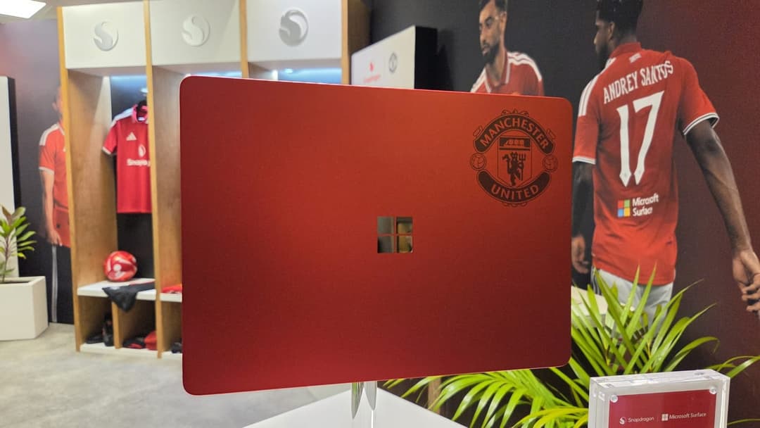 The image shows a red laptop with a Microsoft Windows sticker on the lid, displayed on a white stand in a room with a dark red/maroon wall. The laptop is positioned in front of a wall mural featuring a soccer player wearing a Manchester United jersey with the number 17. The room also contains a white locker room with a