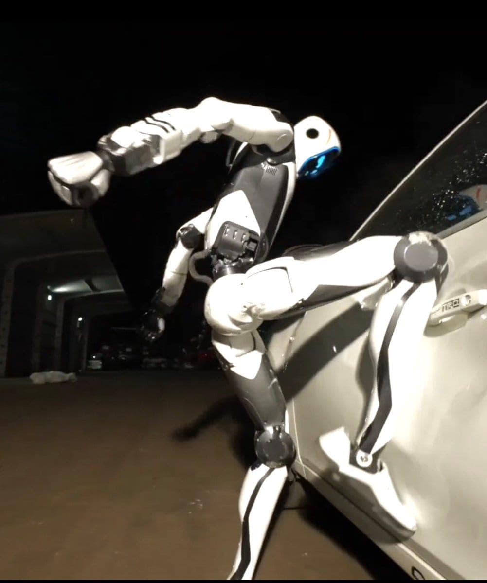 The image shows a robot with a predominantly white and black color scheme, with blue accents on its head and arms. The robot is in a dynamic pose, with its arms and legs extended, suggesting a powerful movement. It is positioned on the right side of the frame, leaning against a white car parked in a dimly lit parking g