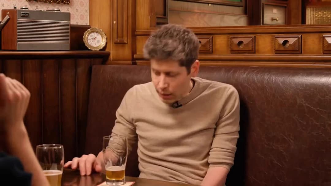 A man in a beige sweater with a small lapel microphone sits in a brown leather booth at a wood-paneled pub table, looking down, with a half-full glass of beer in front of him and another glass partly visible at the left edge. A vintage radio and a small round clock are on a wooden shelf behind him.