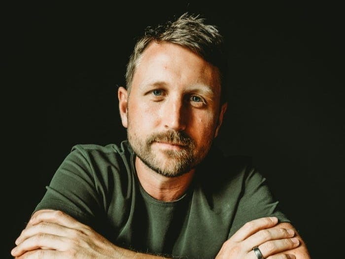 The image shows a man with light brown or blond hair and a beard, wearing a dark green t-shirt. He is seated with his arms crossed, and his gaze is directed towards the camera. He is also wearing a dark-colored ring on his left ring finger. The background is a solid black, which contrasts with the man's green shirt and