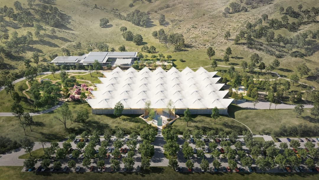 The image presents an aerial view of a modern building with a distinctive white, tent-like roof, nestled amidst a lush landscape. The building is surrounded by a variety of trees and shrubs, and a paved area or parking lot is visible in the foreground. A smaller, separate building with a gray roof is also visible in th